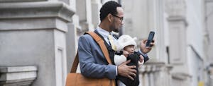 Businessman standing on the street holding his baby in a carrier, looking at his phone