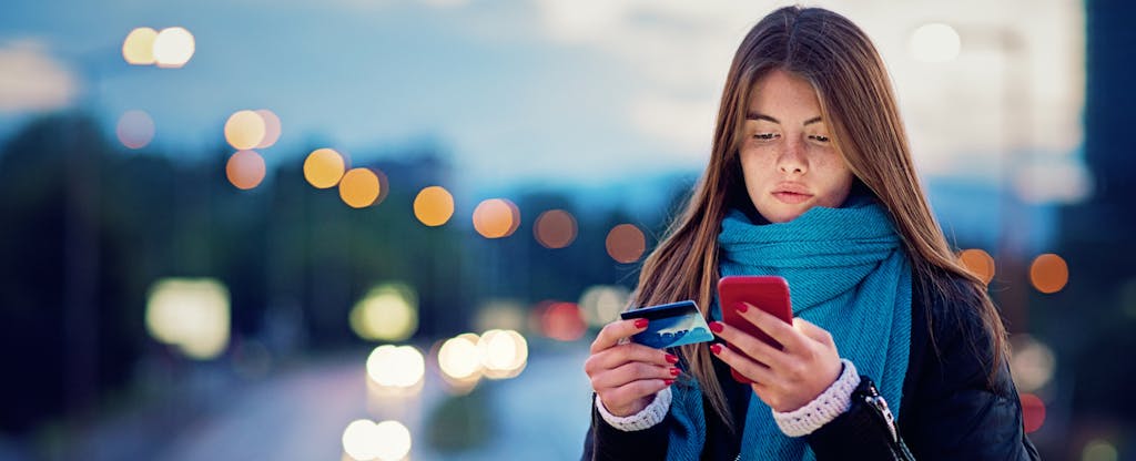 Woman standing outside at night, looking at her phone and credit card