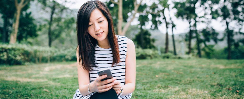 Smiling young woman using smartphone while having picnic and sitting on grassy field