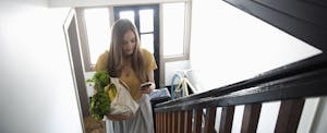 Young woman with smart phone and groceries climbing apartment stairs