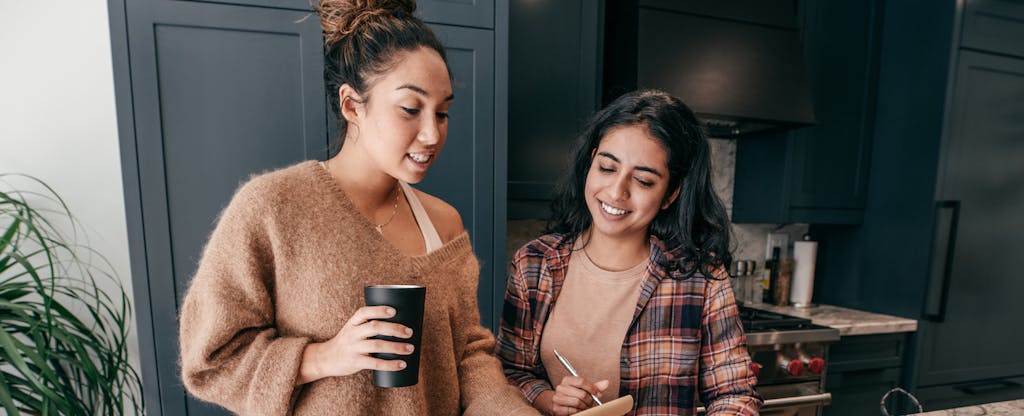 Two young women standing together in their kitchen, having coffee and reading together