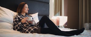 Young woman sitting in bed in hotel room and holding cup of coffee