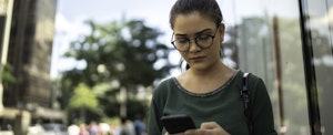 Young woman walking on street, looking at phone and wondering if she can buy a car with no credit