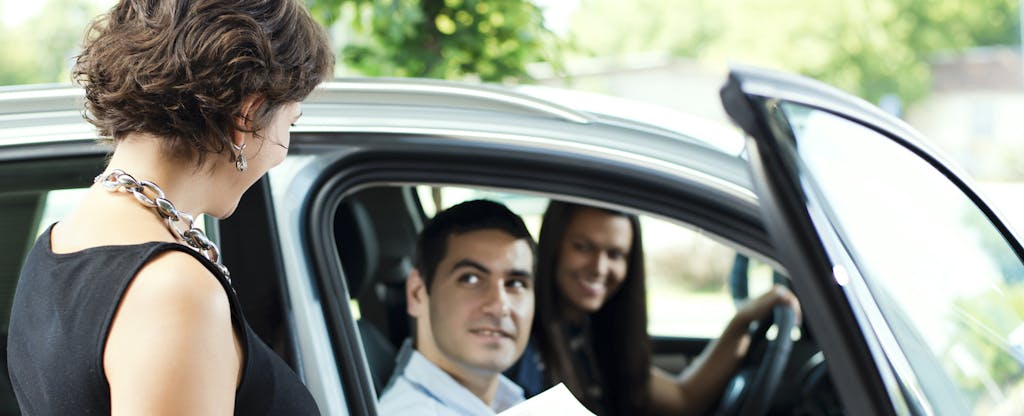 Woman standing outside of a car, holding paperwork and talking to a couple sitting in the car
