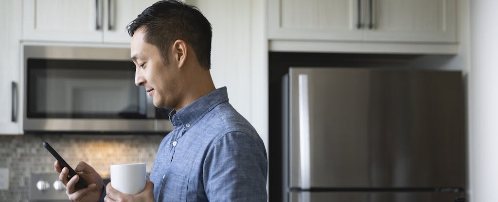 Man standing in his kitchen, holding a cup of coffee and looking at his phone