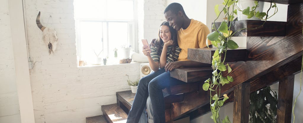 Man and woman sitting together on the stairs in their home, looking at a phone together and smiling