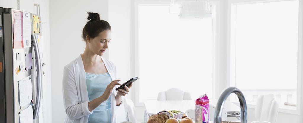 Woman standing in her kitchen, making breakfast and looking at her phone