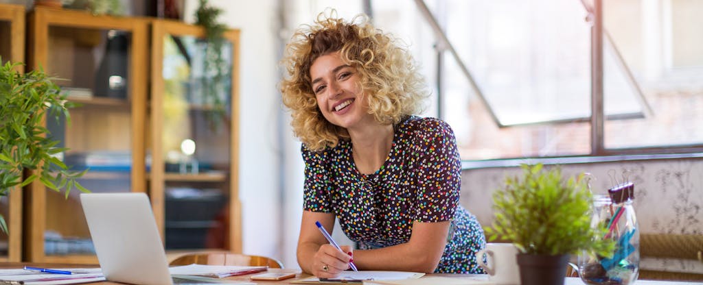 Smiling woman using a standing desk, writing in her notebook with her laptop open