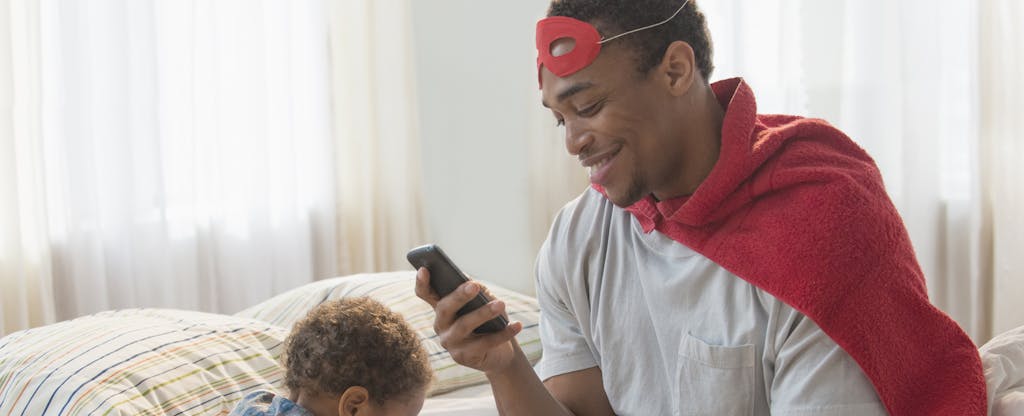 Man playing dress-up with his son, sitting on the bed and checking his phone