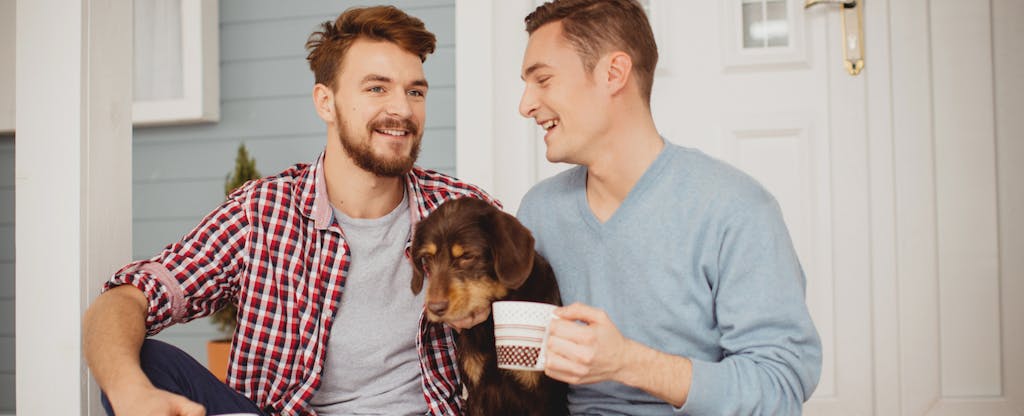 Young same sex couple sitting on porch in front of their home with their dog