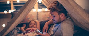 Man sitting in a homemade tent with his young daughter, reading on his phone