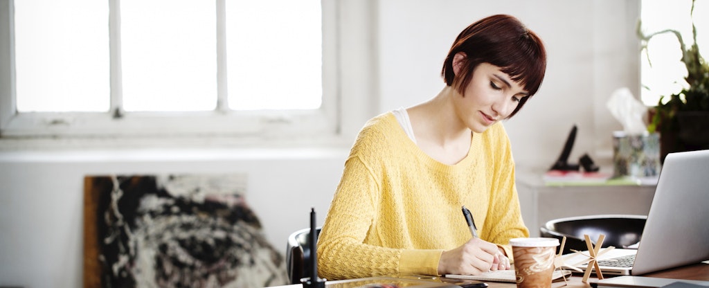 Woman sitting at her desk with her laptop open, writing in a notebook