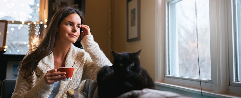 Woman sitting on her couch, holding a cup of coffee and looking out of the window with a thoughtful expression on her face