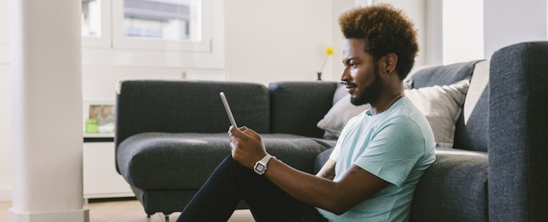 Man sitting on the floor of his living room, leaning against the couch, reading on his phone