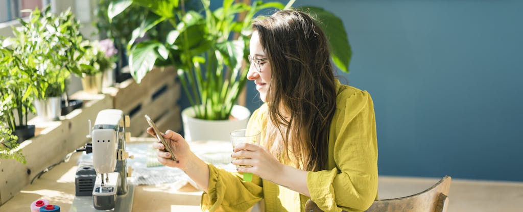 Small-business owner sitting at a desk with a sewing machine, taking a break from work as she reads on her phone