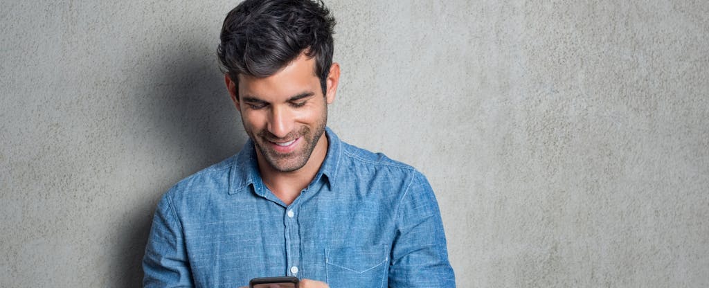 Man standing in front of a gray wall, using his phone and smiling