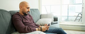 Young man reading text message on his mobile phone while sitting on sofa at home