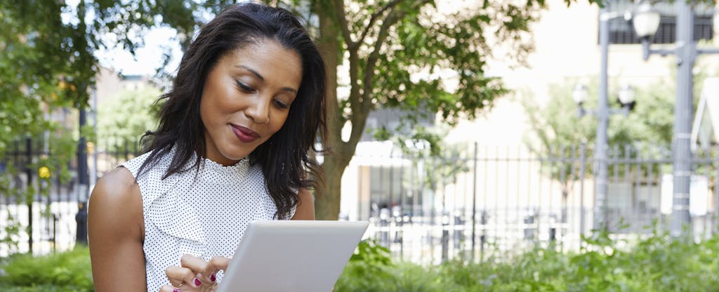 Woman sitting outside in a city park, looking at her tablet and smiling