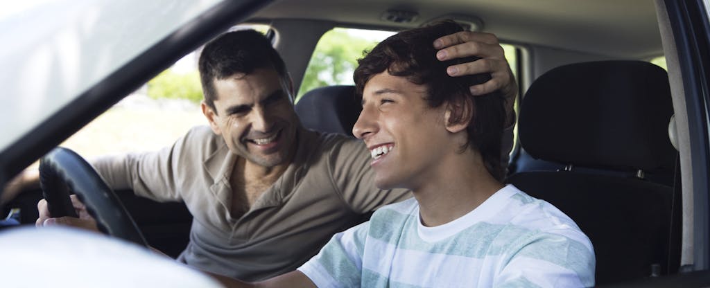 Teenage boy driving a car with his dad in the passenger seat, both of them are smiling
