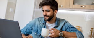 Young man drinking coffee and looking at laptop in his kitchen