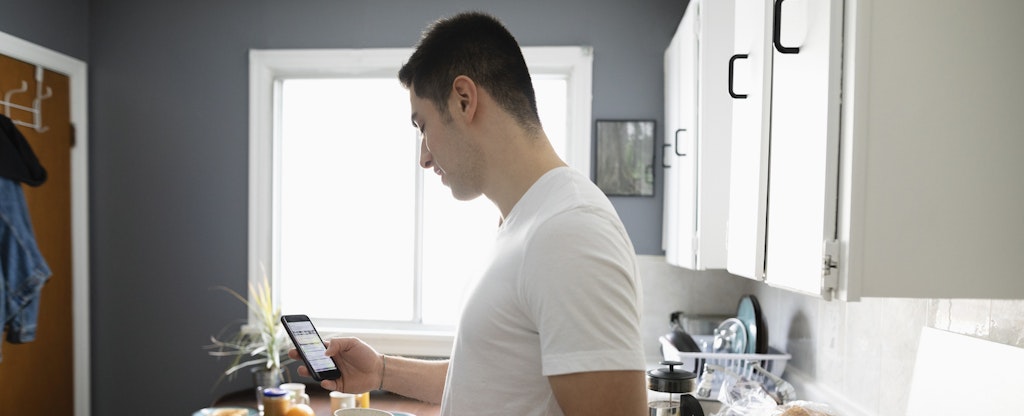 Man standing in his kitchen, drinking coffee and reading on his phone