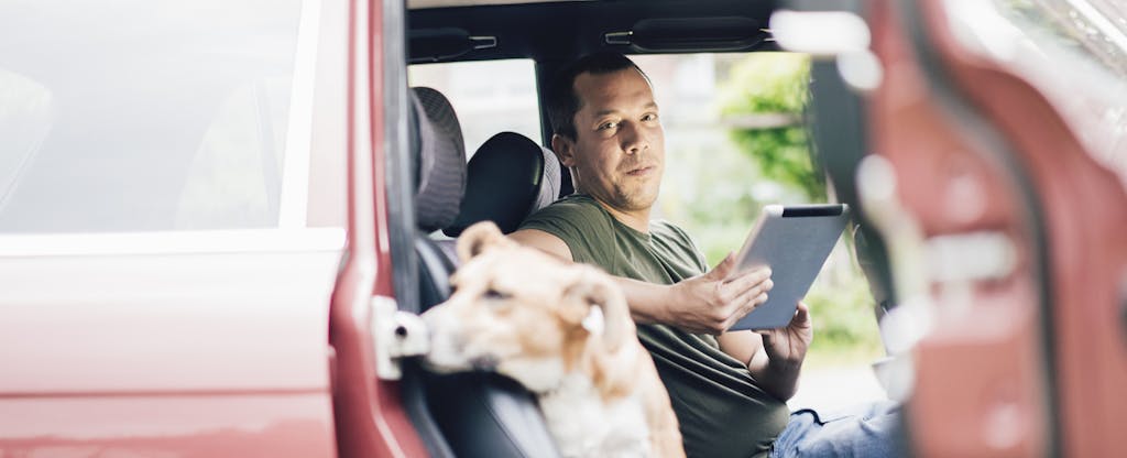 Man sitting in his car, holding a tablet, and looking over at his dog sitting in the passenger seat