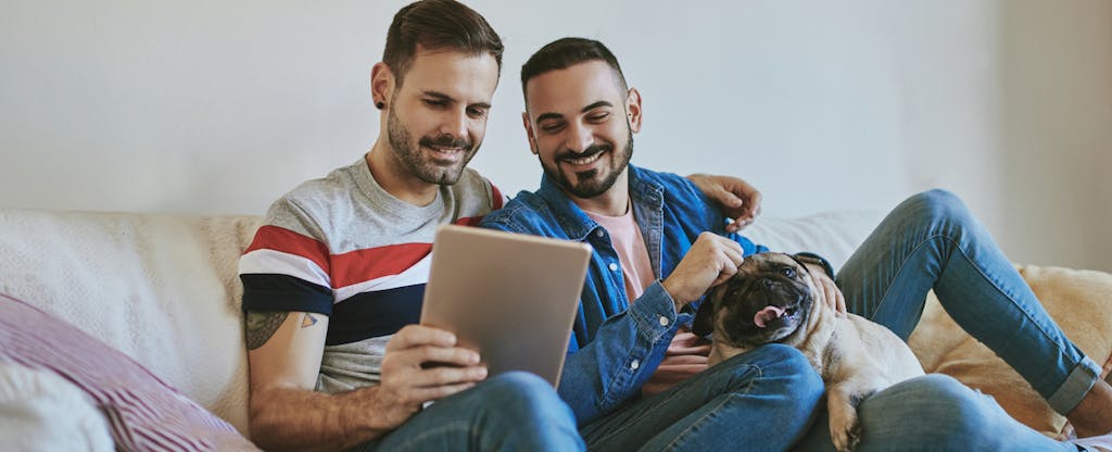 Male same-sex couple sitting on sofa with their dog, using a digital tablet to research the best online loans