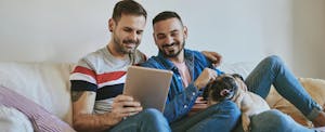 Male same-sex couple sitting on sofa with their dog, using a digital tablet to research the best online loans