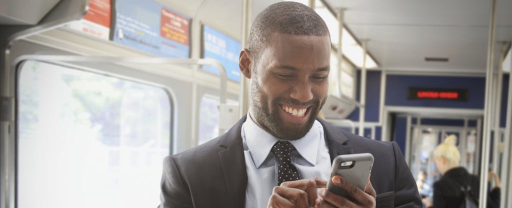 Man standing on a bus, smiling and reading on his smartphone about no-fee banking