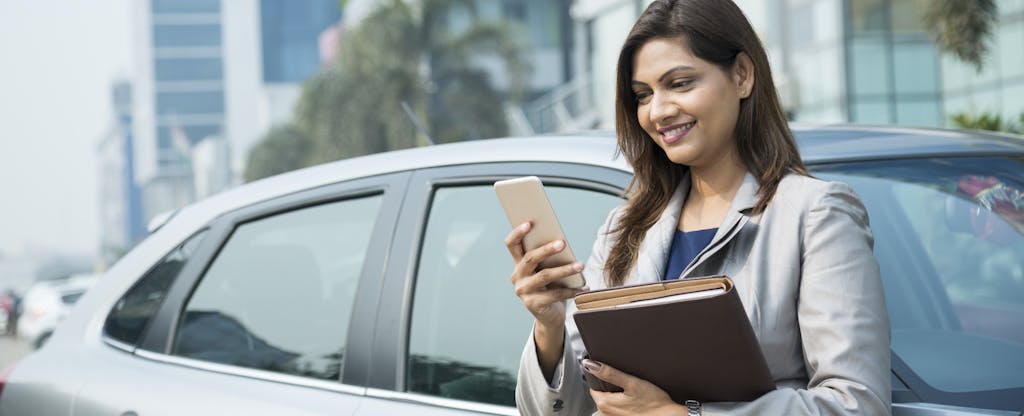 Businesswoman standing next to car with cellphone, looking up rental car insurance coverage on her credit card
