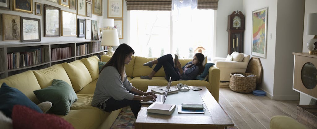 Woman sitting on her couch in her living room, reading about short term capital gains tax on her laptop, with her daughter lying on the nearby couch