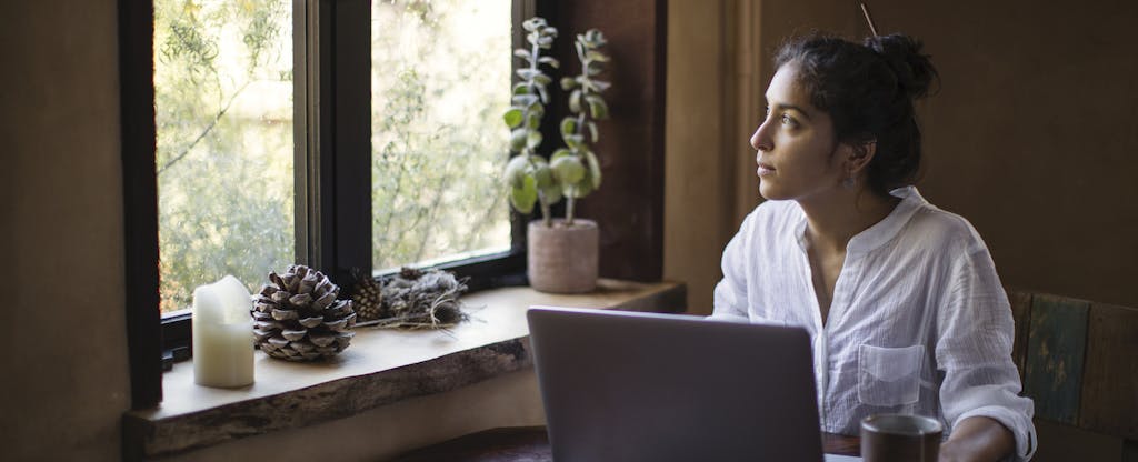 Young woman sitting at table with her computer, looking out the window and wondering when the tax filing deadline is