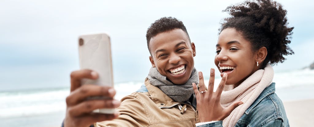 Man and woman standing together on a beach, smiling together for a selfie and showing off the new engagement ring on her hand