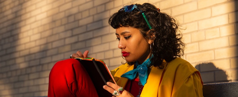 Woman sitting outside, leaning against a wall, writing in her notebook and thinking about getting a National Small Loan