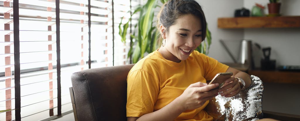 Young woman sitting in a chair at home, looking up where to open a savings account on her phone