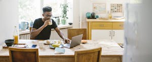 Man sitting at his kitchen table, eating breakfast and reading on his laptop about the best personal loans for fair credit