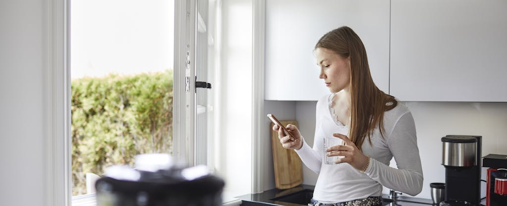 Woman standing in her kitchen, reading on her phone about American Express' Pay It Plan It feature