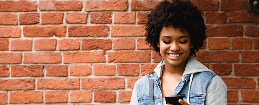 College student leaning against a wall, smiling and reading about savings accounts on her phone