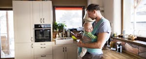 Young father at home in the kitchen holding smart phone, looking up property tax relief, baby son in sling