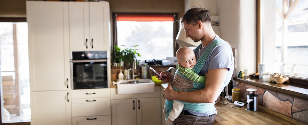 Young father at home in the kitchen holding smart phone, looking up property tax relief, baby son in sling