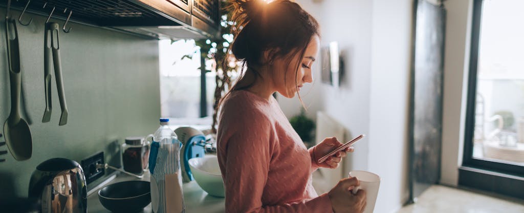 Young woman in her kitchen reading about stimulus check scams on her cellphone
