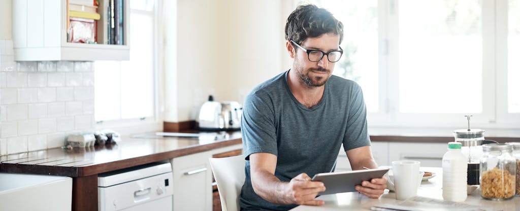 Man sitting in kitchen with digital tablet, looking up trip cancellation insurance
