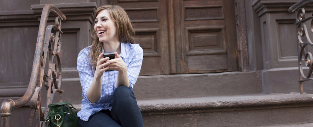 Woman sitting on the steps outside her apartment, smiling as she uses the Brigit app on her phone