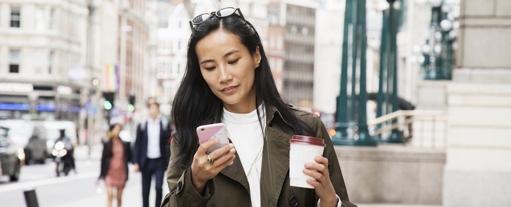 Woman walking on city street with coffee, looking up eos cca credit reports on her cellphone