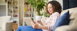 Woman sitting on sofa drinking coffee, looking up neobanks on her digital tablet