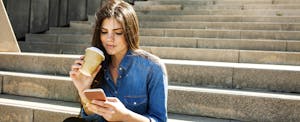 Young woman with coffee sitting outside on stairs, using cellphone to look up current balance vs available balance