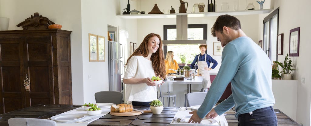 A young couple in the background, cooking dinner in their new home, while their friends set the kitchen table.