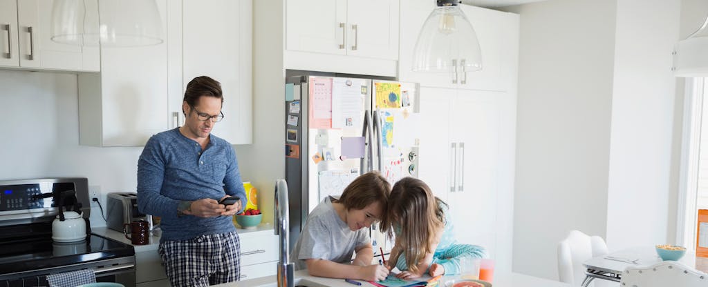 Father texting while children color in kitchen in the morning