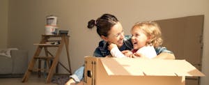Laughing woman with daughter sitting in cardboard box in their new home they are moving into