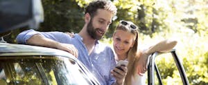Smiling young couple with cellphone standing by car in the woods
